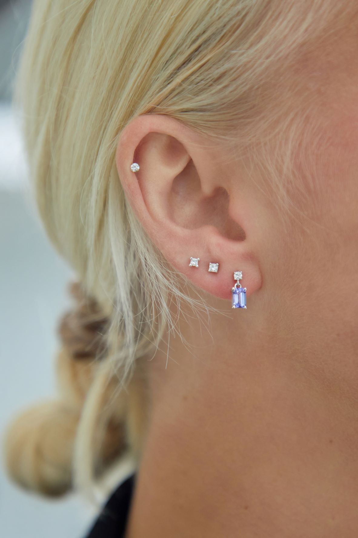 Close-up of a person's ear wearing multiple stud earrings, including a small silver hoop in the cartilage, two small square stud earrings, and a dangling silver earring with a blue and purple crystal drop.