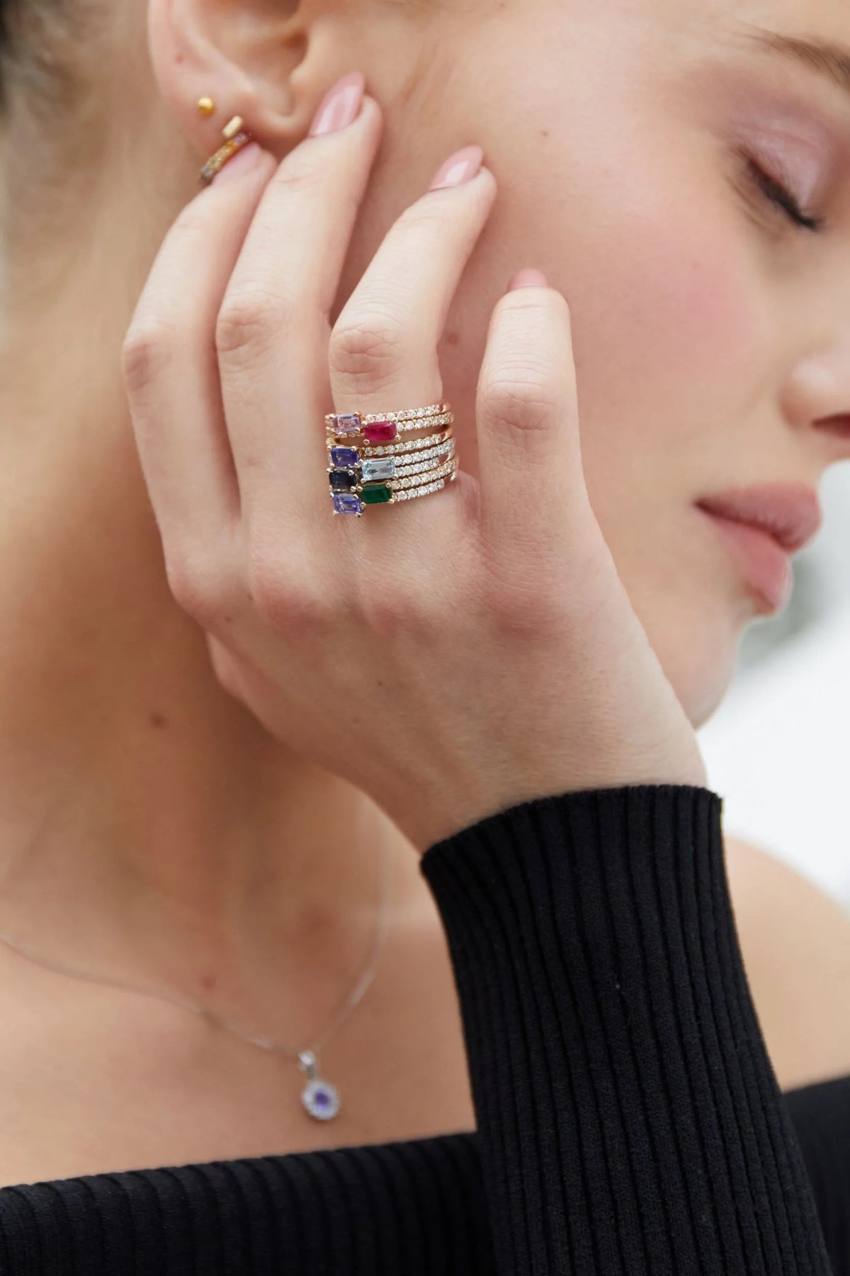 Close-up of a woman's hand wearing multiple rings with colorful gemstones, against a blurred background.